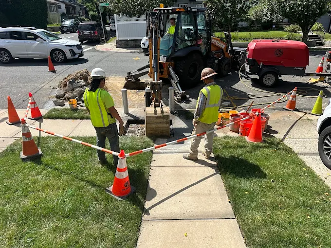 Construction workers repairing sidewalk with heavy machinery, orange cones, and safety barriers on a sunny day.